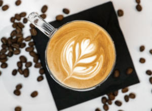 A cup of coffee with a cream flower at the top of the glass. The glass is surrounded by coffee beans.